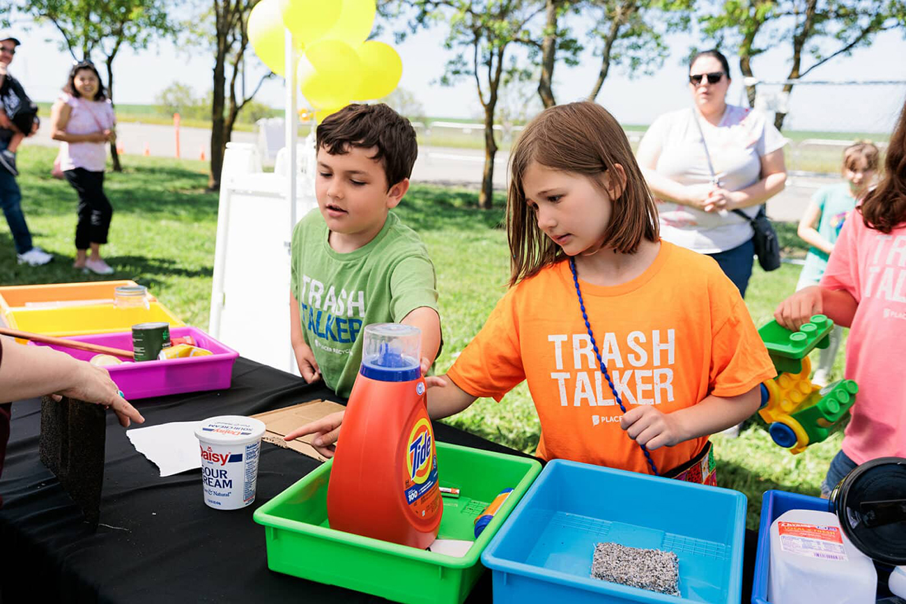 Kids wearing "Trash Talker" T-shirts examine recycling bins filled with household containers at an outdoor event.