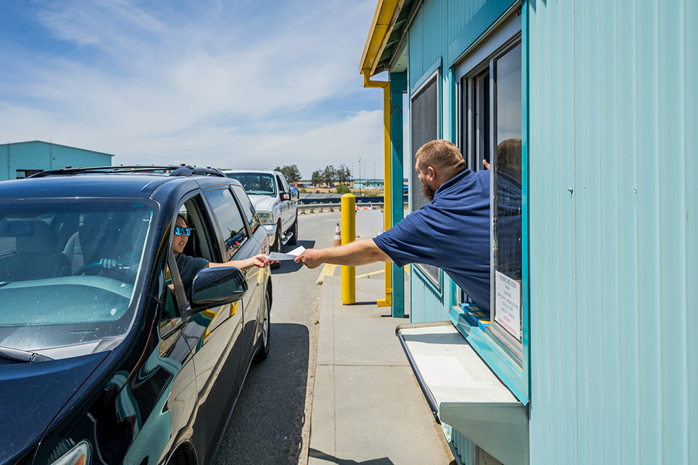 A man in a blue shirt leans out of a turquoise booth window, handing a ticket to a driver in a black SUV. The sky is clear and sunny.