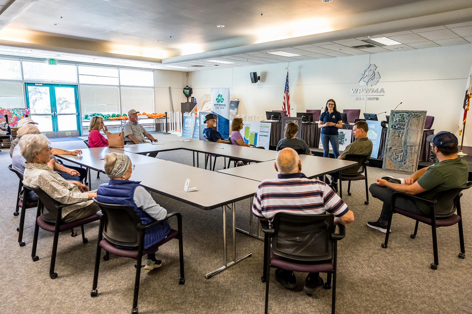 A group of people seated around a large table in a WPWMA meeting room, listening to a speaker.