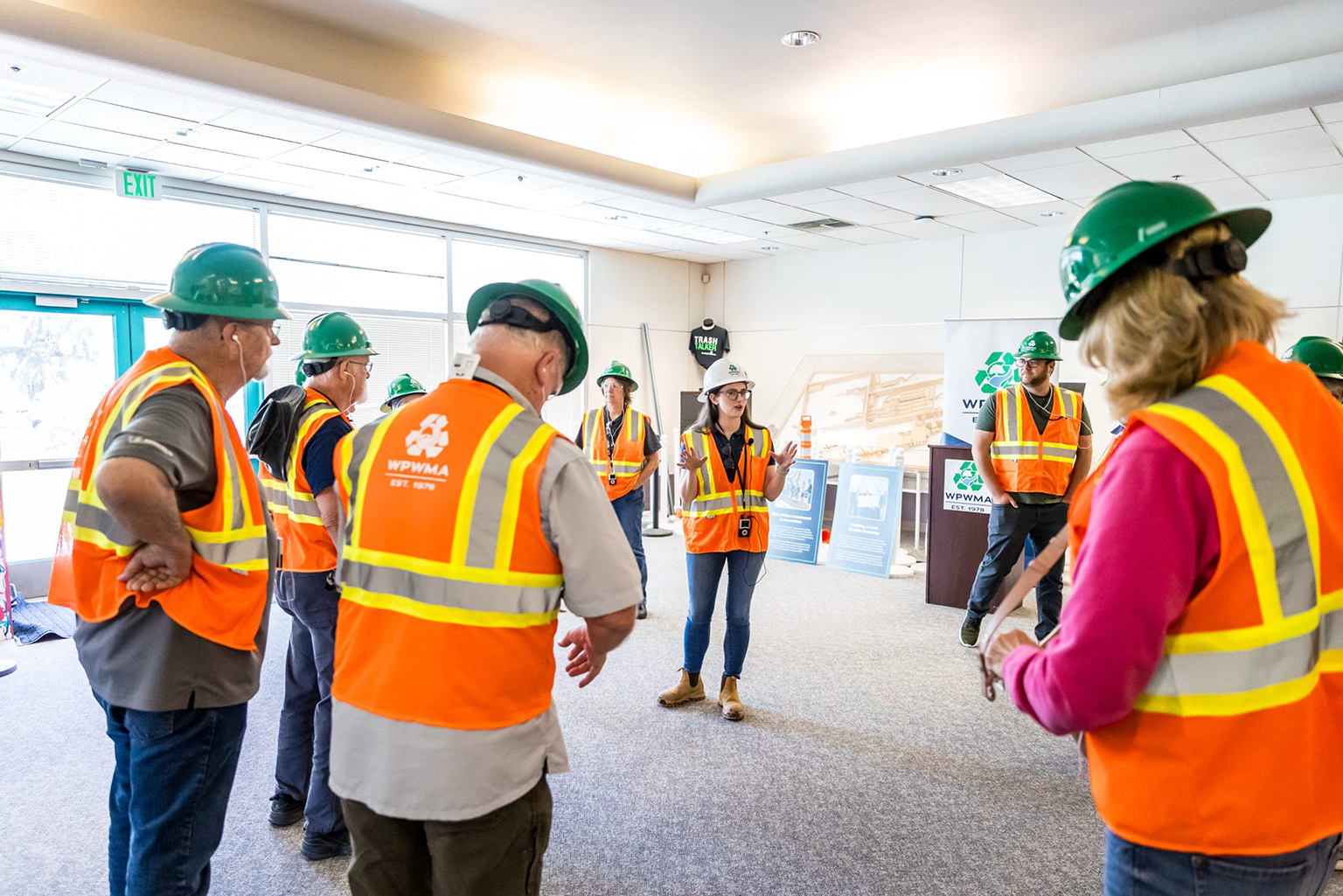 A group of people in safety vests and hard hats are gathered indoors, listening to a speaker in a briefing for the WPWMA MRF tour