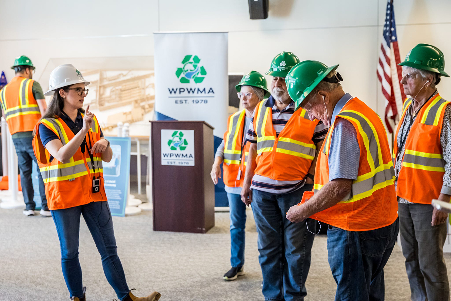 A group of people in safety vests and hard hats listens to a speaker in a conference room with a WPWMA banner.