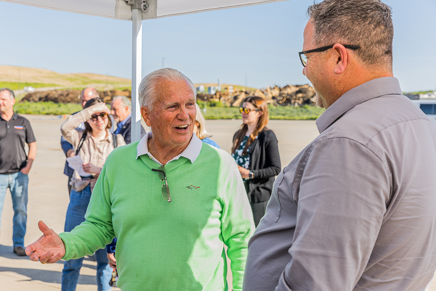 A mature man in a bright green sweater conversing with another man in glasses and gray shirt
