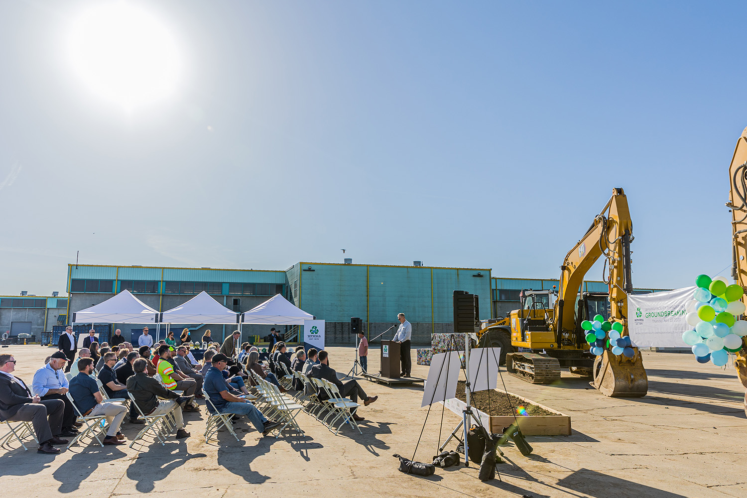 A crowd gathers outdoors for a ceremony, featuring a speaker at a podium, excavators, and tents under a clear sky.