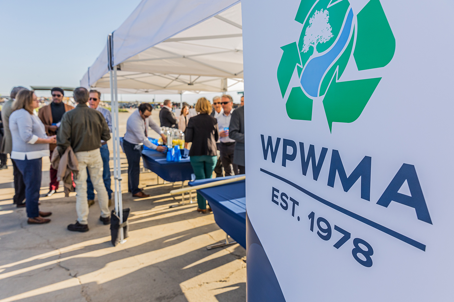 A group of people mingles under a tent at an outdoor event, with a sign displaying "WPWMA EST. 1978" in the foreground.
