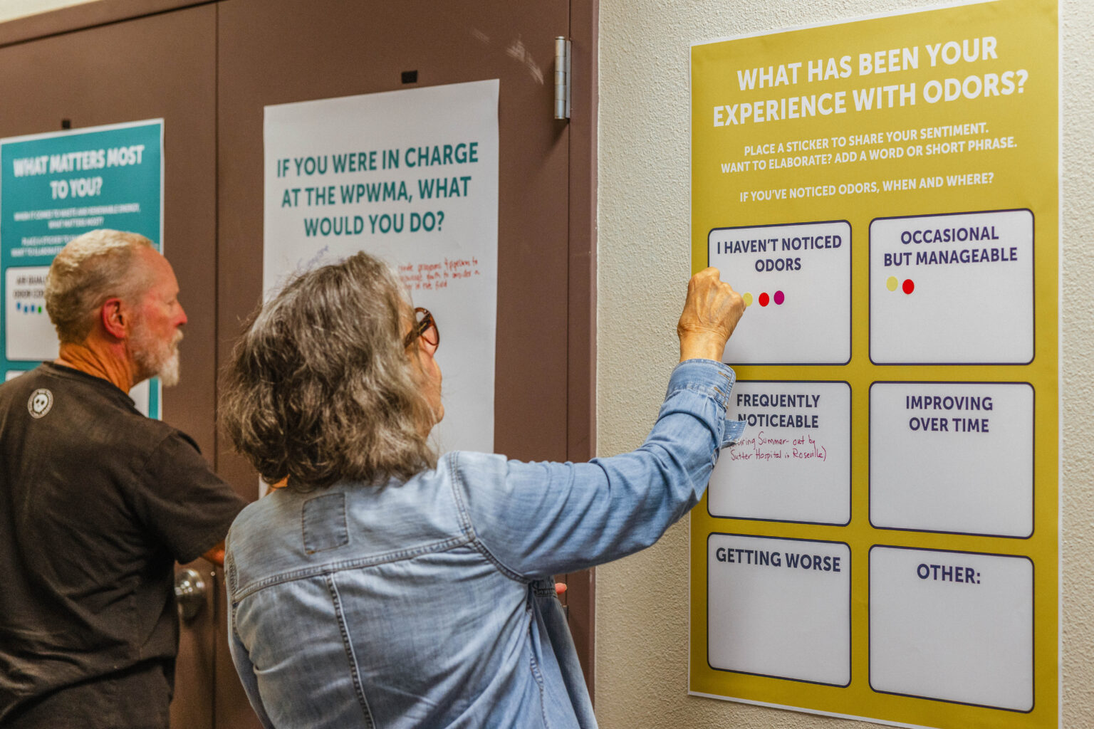 Man and woman write sentiments and place stickers sharing sentiments on printed posters asking attendees questions like "What matters most to you?", "If you were in charge at the WPWMA, what would you do?" and "What has been your experience with odors?"