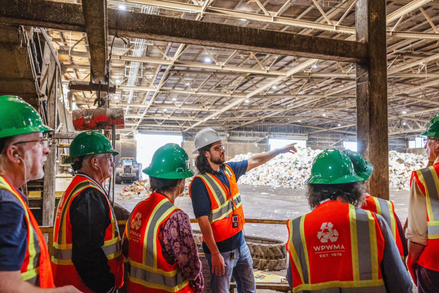 Man leads tour through recycling facility with attendees and tour guide all wearing hard hats and orange safety vests