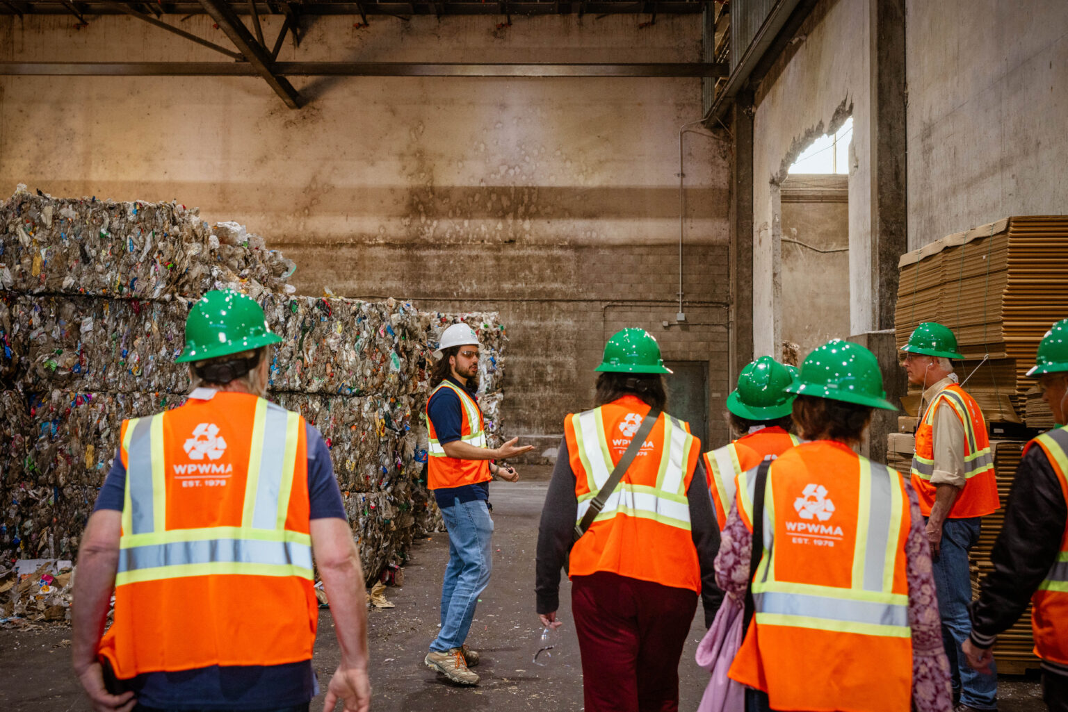 Man leads tour through recycling facility with attendees and tour guide all wearing hard hats and orange safety vests