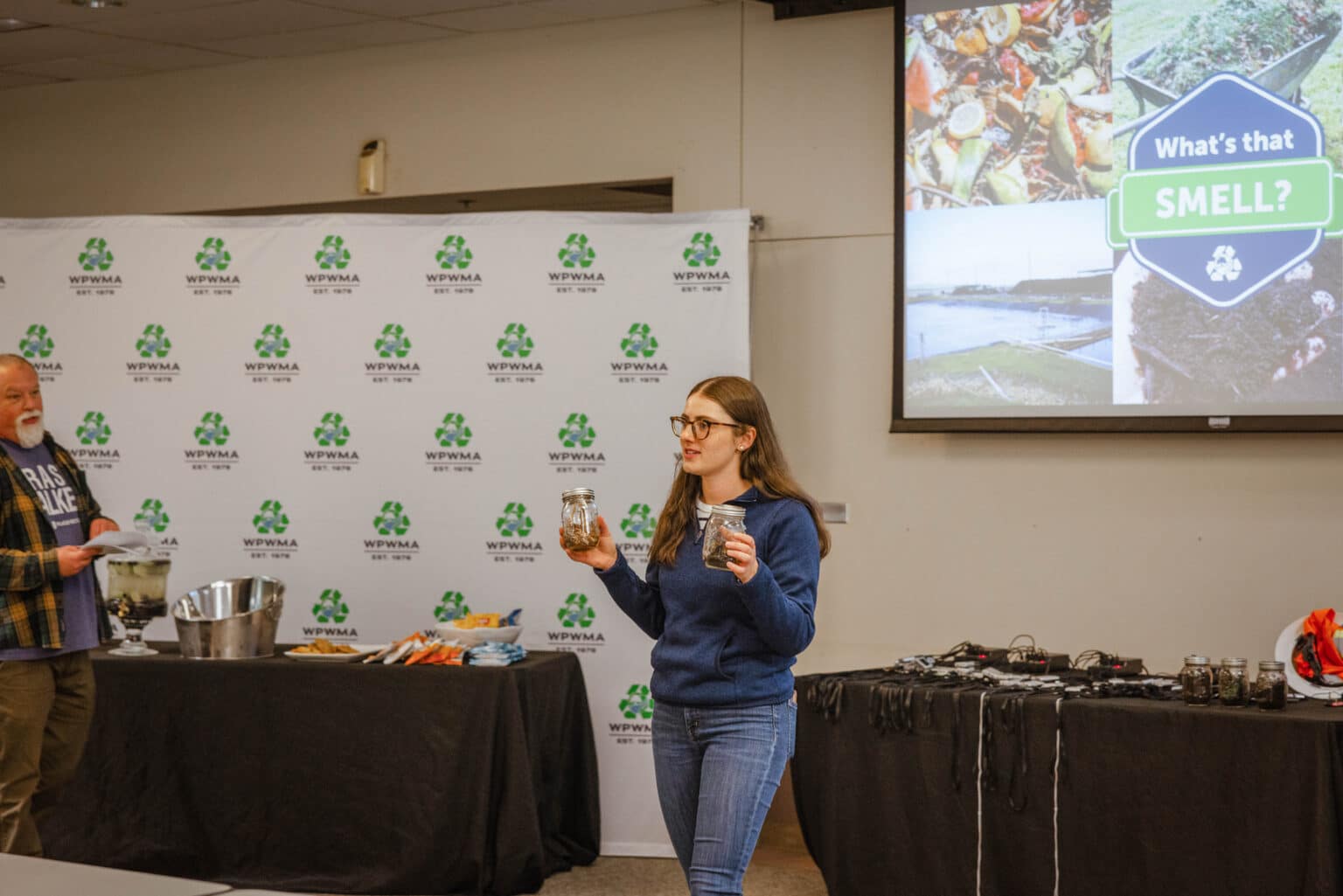 Woman holds jars of materials in front of a PowerPoint slide that says "What's That Smell" with a variety of photos of odor sources from the WPWMA's campus