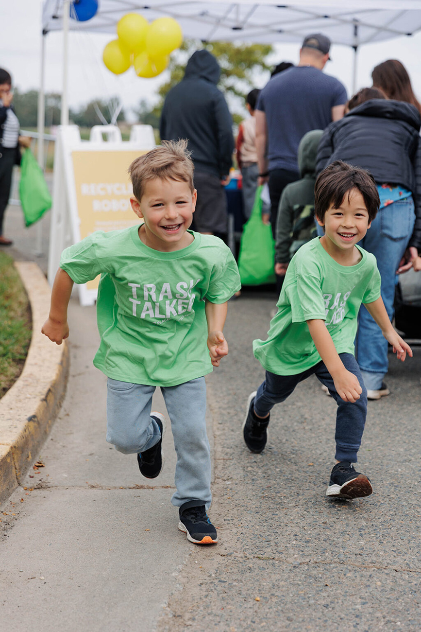 Two happy boys smiling and running with matching light green shirts that say "TRASH TALKERS" near an event booth at the WPWMA Trash Bash event