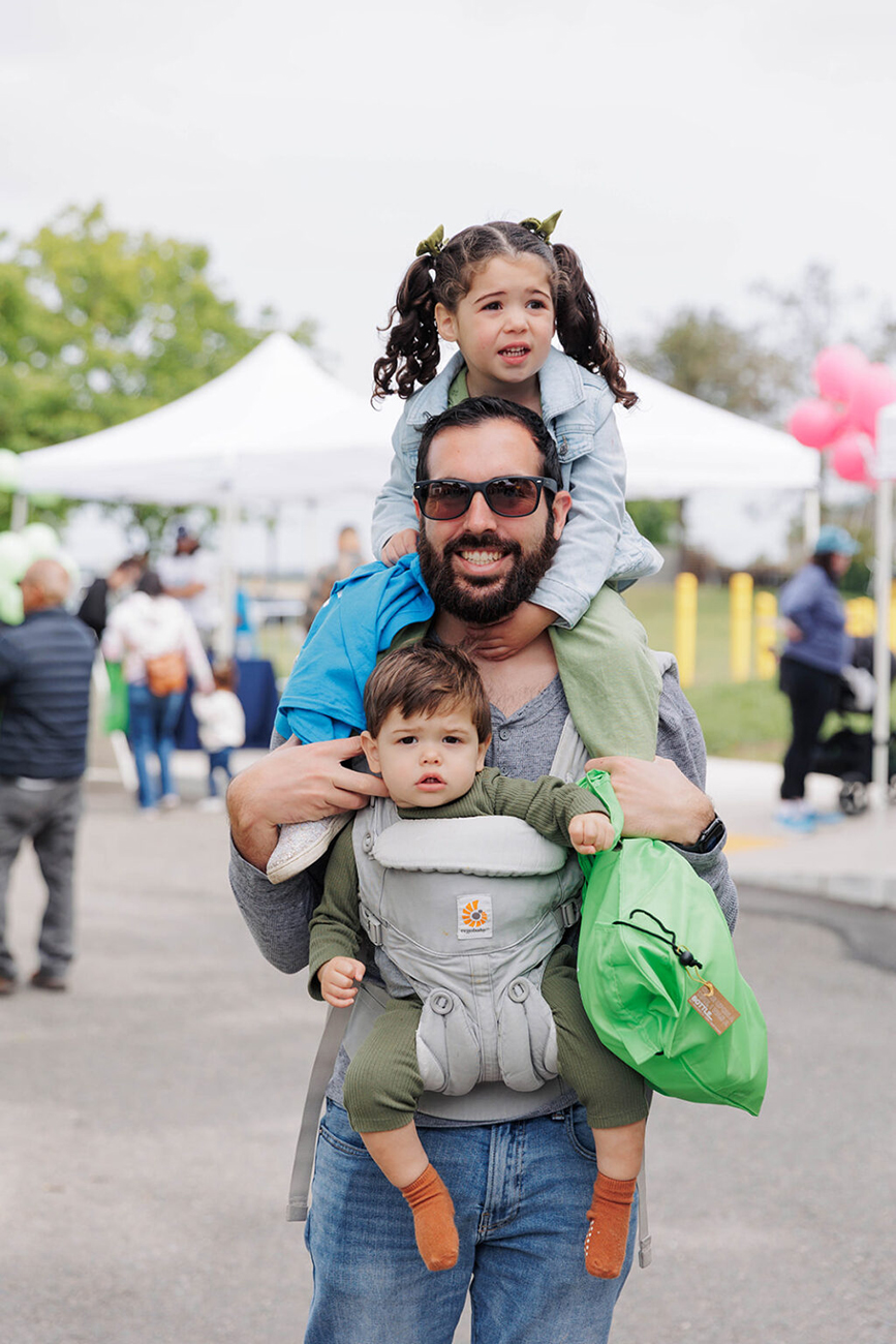 A man wearing sunglasses smiles while carrying a young girl with pig tails on his shoulders and a baby boy in a grey front-facing carrier behind them are event booths at the WPWMA Trash Bash event