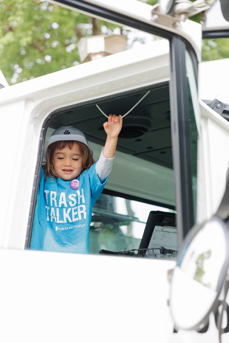 A young child inside a white truck wearing a sky blue "Trash Talker" shirt from the WPWMA Trash Bash event