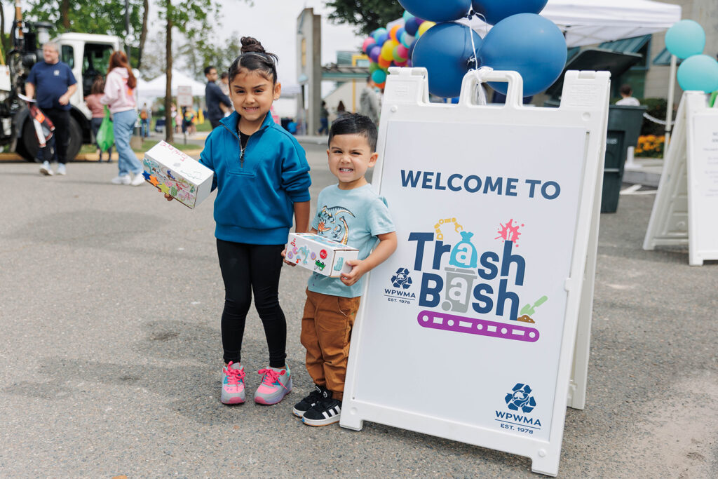 Two kids hold decorated boxes beside a "Welcome to Trash Bash" sign from WPWMA.