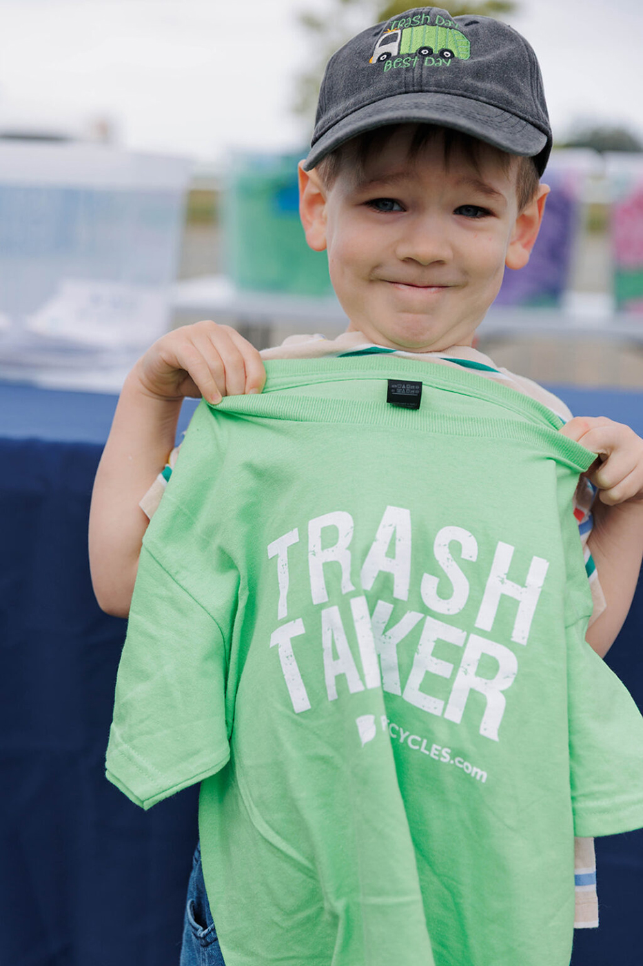 A smiling boy holding up a light green crewneck t-shirt with "TRASH TALKER" printed in white on the front during the WPWMA Trash Bash event