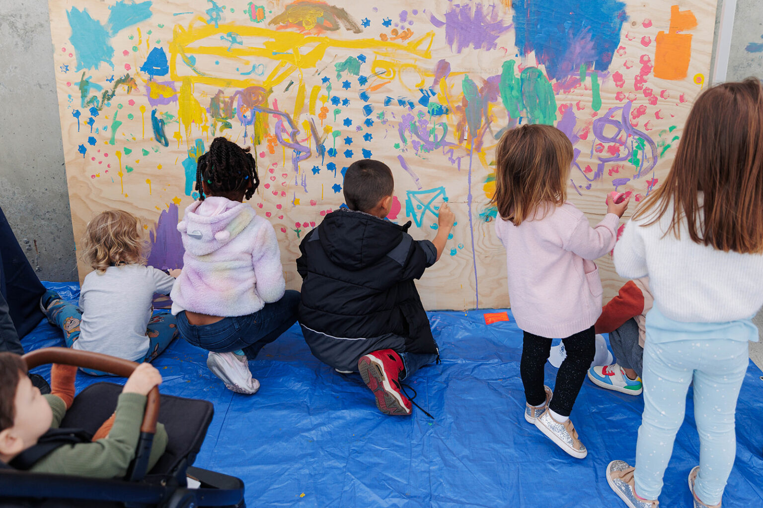 Young children are seen from behind as they paint a large wooden board attached to a wall. They use vibrant, multi-colored paints, adding to a busy abstract mural. The children are sitting and standing on a blue tarp spread across the floor to catch any drips. In the foreground, another child sits in a stroller, looking toward the mural