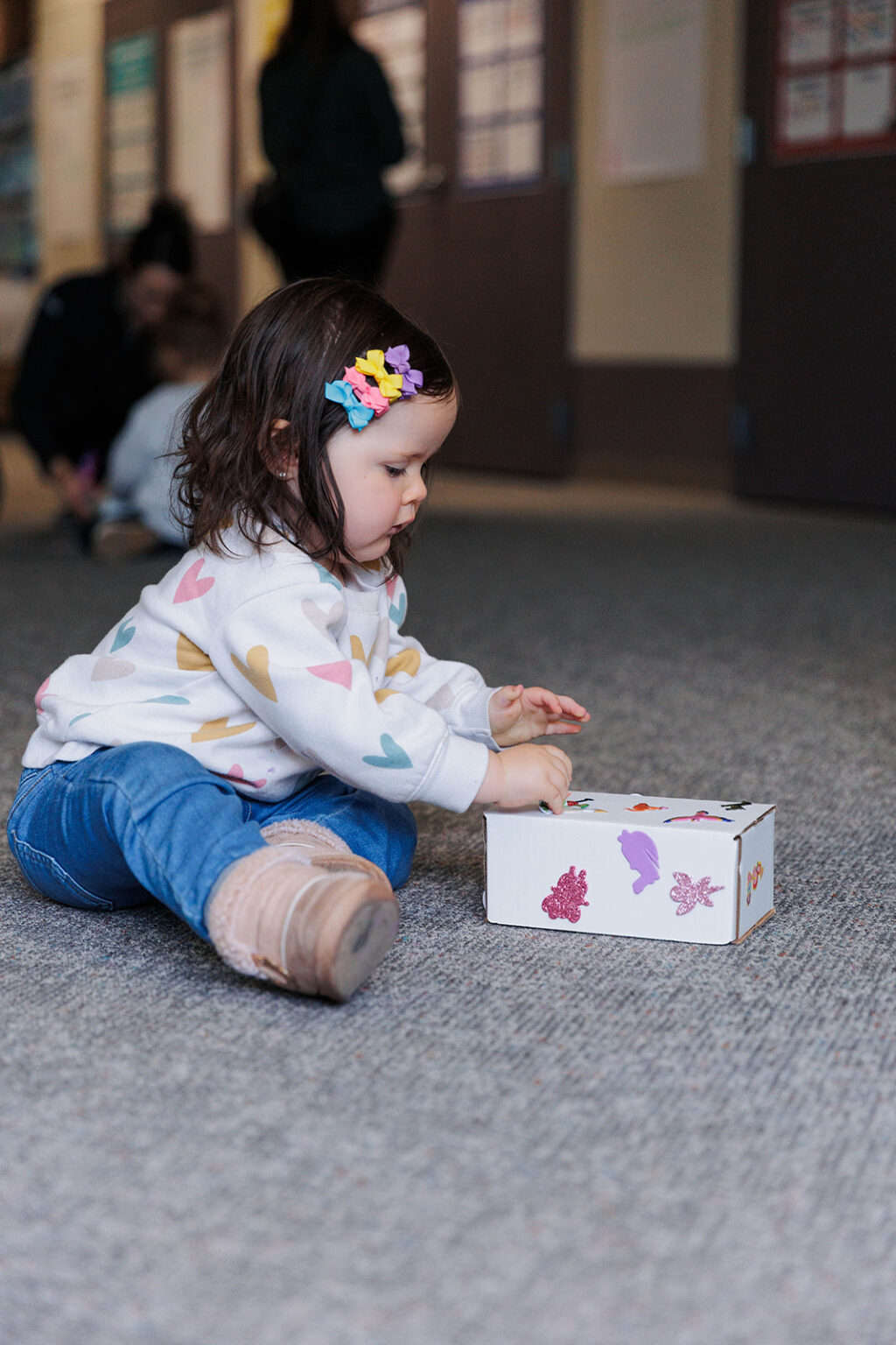 A female toddler with colorful ribbon clips on her hair engaging in a learning activity with a decorated box while sitting on the floor
