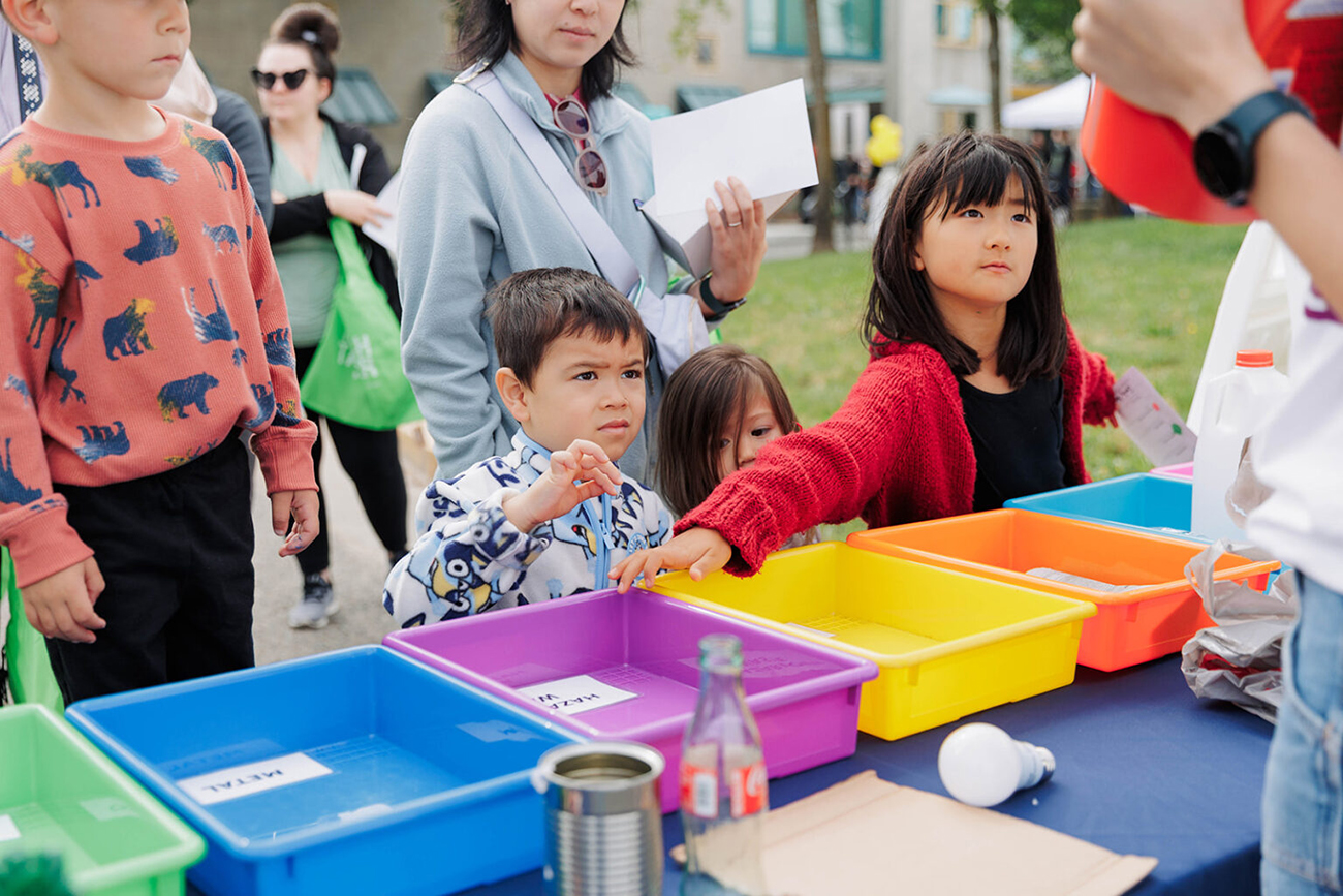 A group of children standing in front of a table with multicolored bins outdoors to participate in a trash sorting game at the WPWMA's Trash Bash event
