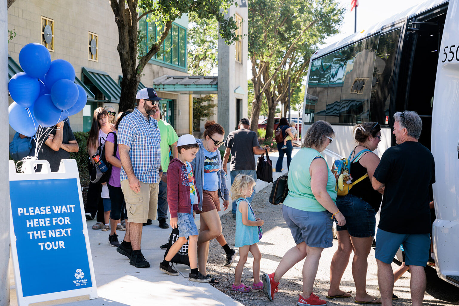Group of people, both adults and young children are lined up outdoors to board a large white bus to tour the WPWMA Facility in California during the Trash Bash event