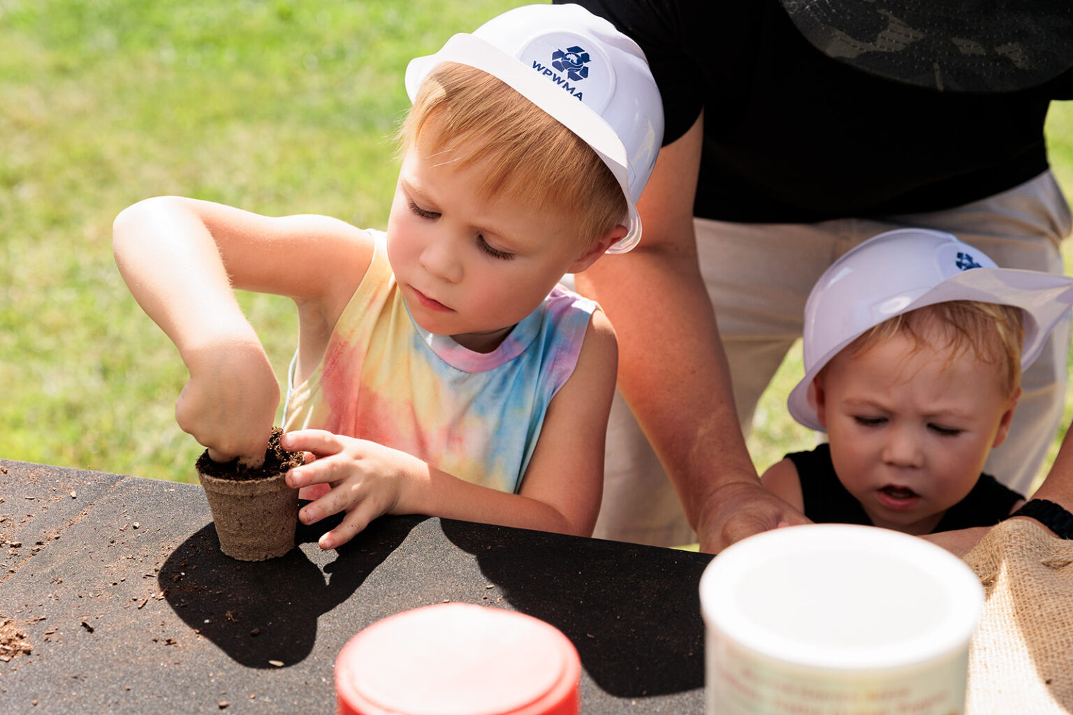 Two young children are planting in small pots at a table, wearing white hats shaped like a hard hat with WPWMA logo. One child is focused on digging in the soil.