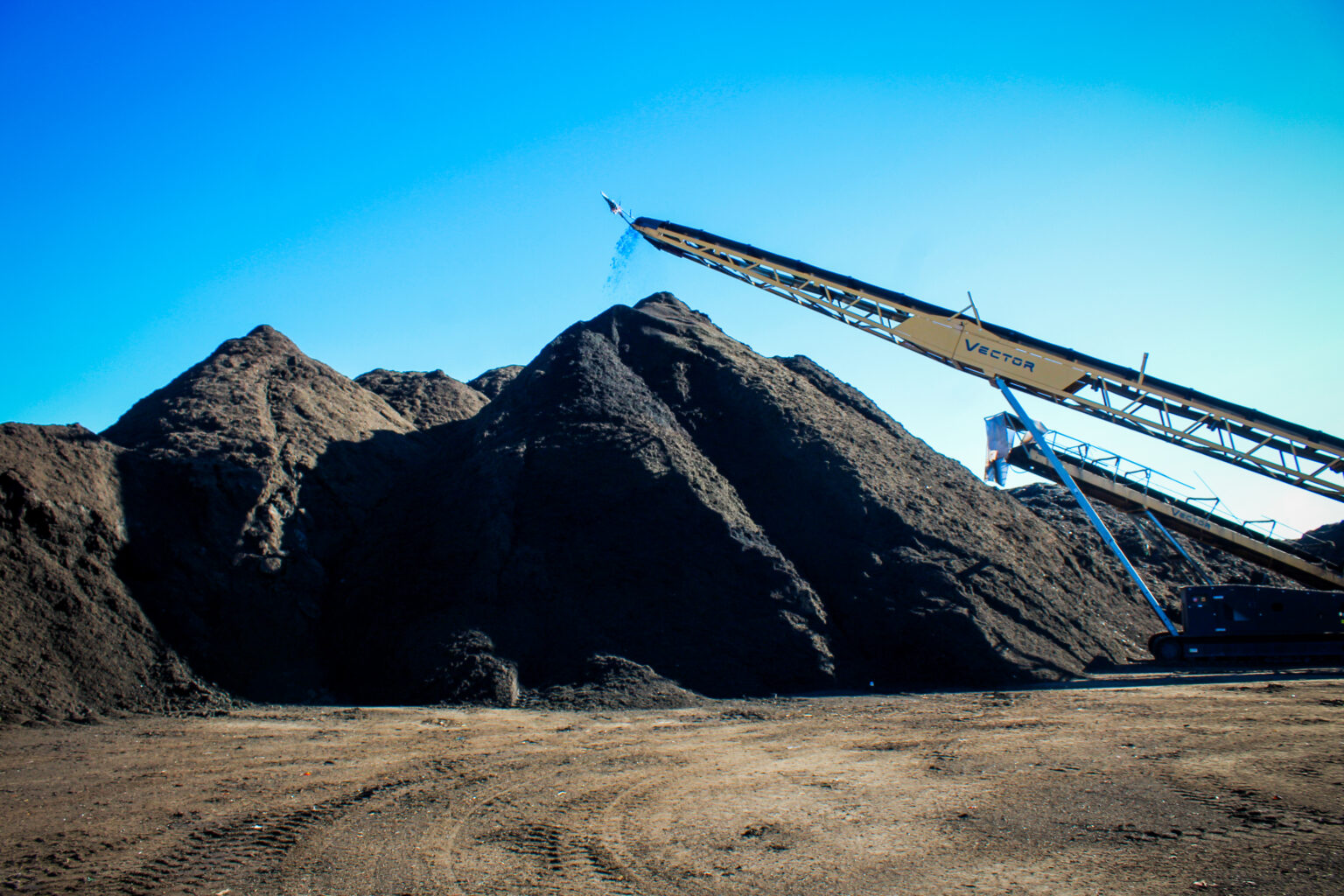 A conveyor belt stacks large piles of finished compost under a clear blue sky