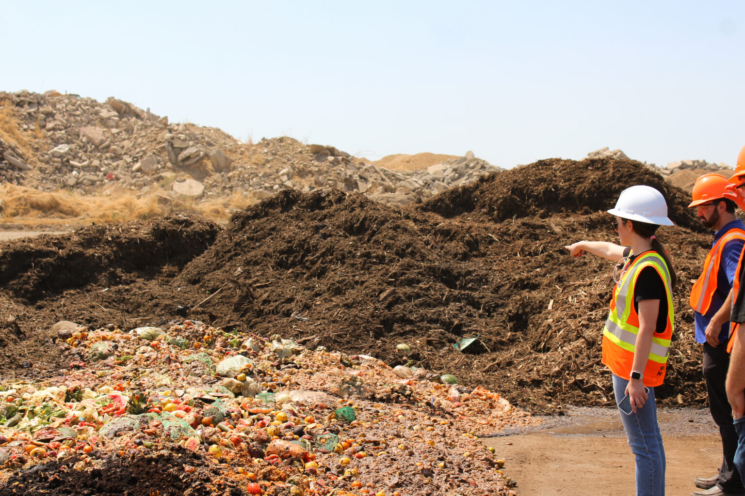 Woman in a white hard hat and safety vest pointing at dark brown organic waste windrow piles with fresh heap of multicolored food scraps and organic waste in an outdoor composting facility under a clear blue sky