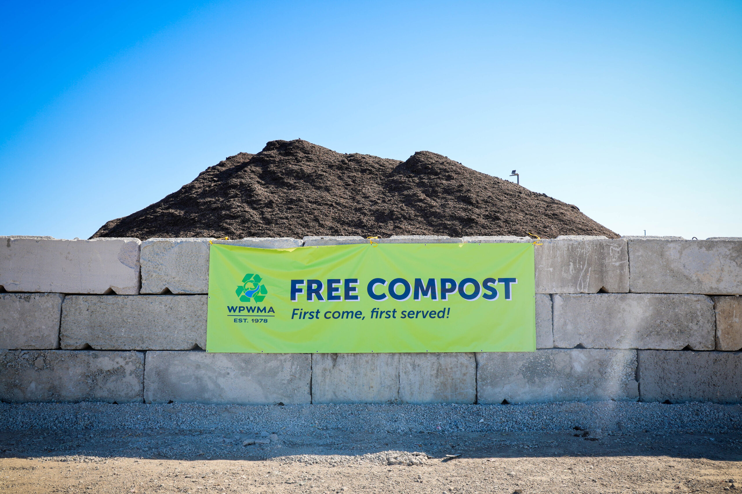 Concrete bunker with overflowing brown compost soil with a green banner that says, "FREE COMPOST"