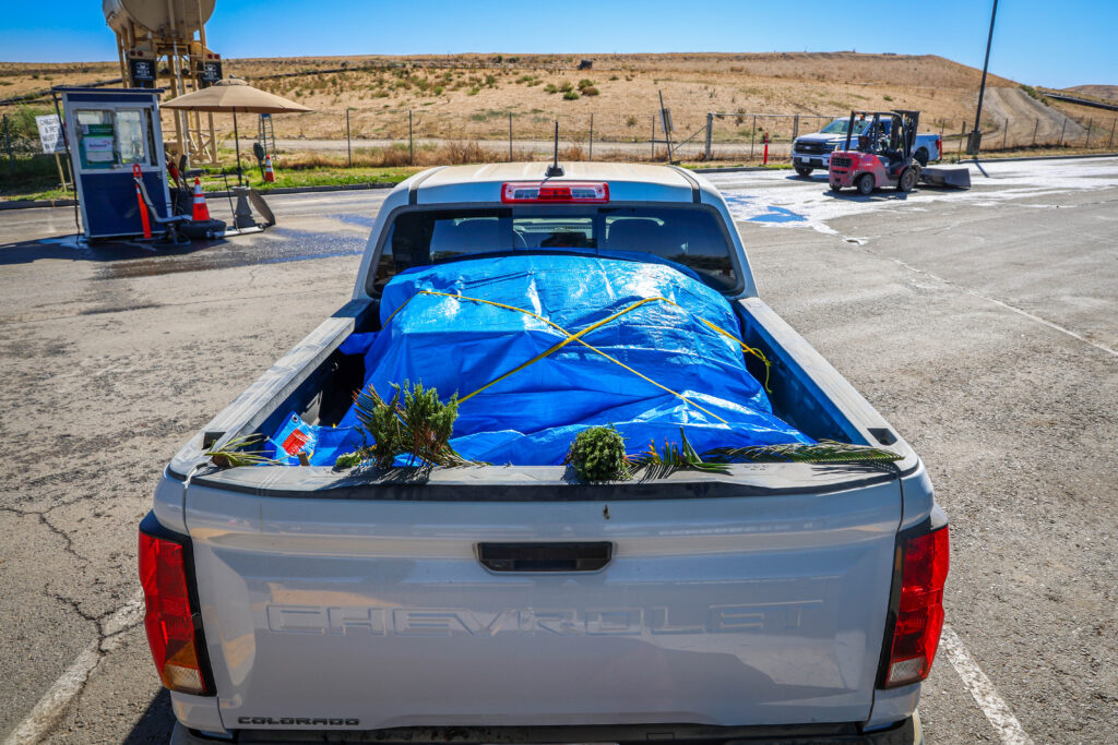 White truck with a bed full of green waste covered with a blue tarp and tied down securely with a yellow rope